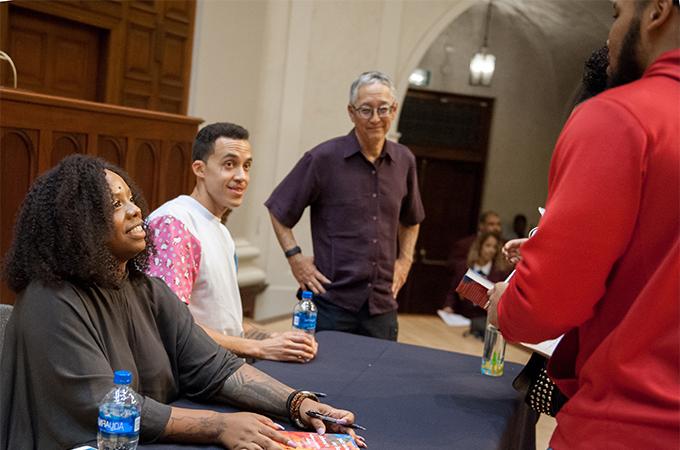 Speakers sit at a table to sign books while greeting members of the audience.